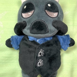 May include: A plush toy creature with a grey head, large eyes, and small white horns. It wears a blue shirt and a black vest with decorative buttons. The toy is approximately 15 cm tall.