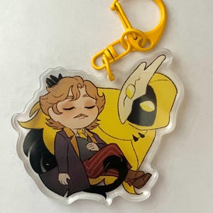 Puede incluir: Un personaje de dibujos animados amarillo y negro con una corona y un bigote está sentado sobre un pájaro amarillo y negro con alas. El personaje lleva un abrigo morado, pantalones a rayas rojas y blancas y zapatos marrones. El personaje tiene los ojos cerrados y está durmiendo. El personaje y el pájaro están en un llavero de acrílico transparente con un anillo de metal amarillo.