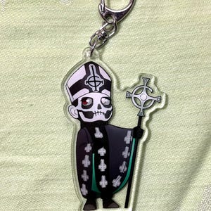 May include: A clear acrylic keychain featuring a cartoon character in a black and white outfit with a skull face and a tall hat. The character is holding a silver cross and has a silver keychain attachment.