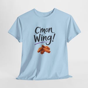 Op de afbeelding: Lichtblauw T-shirt met de tekst "C'mon Wing!" in zwarte letters, onderstreept met paars, en een afbeelding van gefrituurde kippenvleugels. Het T-shirt heeft een ronde hals en korte mouwen.