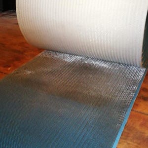 Puede incluir: Un rollo de tela blanca texturizada está parcialmente desenrollado, revelando un material azul oscuro texturizado debajo. Los materiales parecen estar en capas, con la tela blanca en la parte superior. La imagen está tomada sobre una superficie de madera.