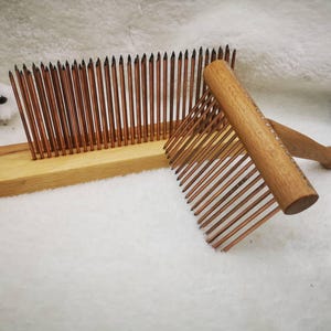 Könnte beinhalten: Zwei Wollkämme aus Holz. Einer ist ein stationärer Kamm mit vielen dünnen, kupferfarbenen Zinken, die in einer Holzbasis befestigt sind. Der andere ist ein Handkamm mit Holzgriff und kupferfarbenen Zinken. Die Kämme dienen zur Faseraufbereitung.