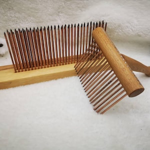 Könnte beinhalten: Zwei Wollkämme aus Holz. Einer ist ein stationärer Kamm mit vielen dünnen, kupferfarbenen Zinken, die in einer Holzbasis befestigt sind. Der andere ist ein Handkamm mit einem Holzgriff und kupferfarbenen Zinken. Die Kämme dienen zur Faseraufbereitung.