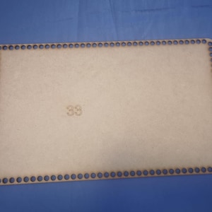 Può includere: Una base rettangolare in legno con fori distanziati uniformemente lungo il perimetro. La base è di colore beige chiaro, con il numero "33" stampato al centro. Lo sfondo è blu unito.