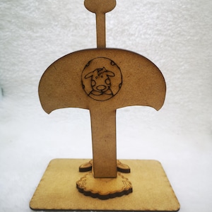 Puede incluir: Un soporte decorativo de madera con una ilustración de vaca dentro de un marco circular. El soporte tiene una base rectangular, un soporte central y una parte superior redondeada. La ilustración de la vaca está en el centro de la parte superior en forma de paraguas.