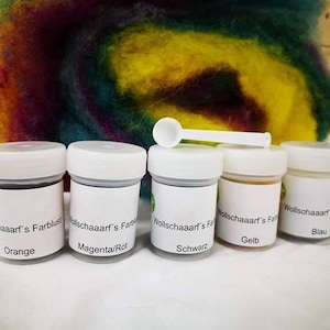 Könnte beinhalten: Fünf kleine weiße Gläser mit Etiketten, die verschiedene Farbstoffe enthalten. Die Etiketten lauten "Orange", "Magenta/Rot", "Schwarz", "Gelb" und "Blau". Ein kleiner weißer Löffel liegt auf den Gläsern, mit einem bunten Wollhintergrund.