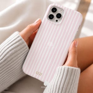 May include: A smartphone case featuring a vertical light pink and white striped pattern. The case has a cutout for a triple-lens camera system on the back. The letters ADJ are printed in a gold-colored serif font near the bottom of the case.