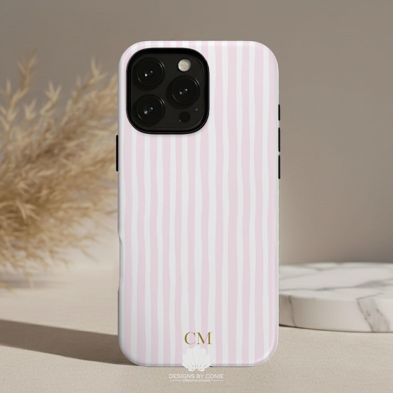 May include: A phone case with a pink and white striped pattern. The case has a black camera module and black side buttons. The bottom of the case has the initials "CM" and the text "DESIGNS BY CONIE GRAPHIC STUDIO".