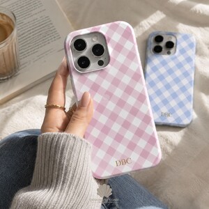 May include: A pink and white gingham patterned phone case with the letters DBC printed in gold near the bottom. A second phone case with a blue and white gingham pattern and the letters DBC is visible in the background.