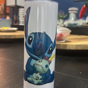 May include: A white tumbler featuring a blue cartoon character, holding a doll. The character has large eyes and pointed ears. The tumbler includes a clear straw and a lid. The design is a popular character from a well-known animated film.