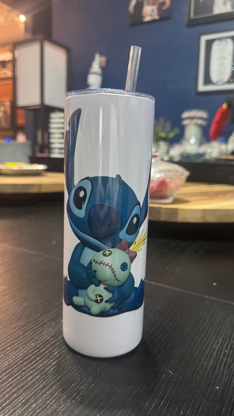 May include: A white tumbler featuring a blue cartoon character, holding a doll. The character has large eyes and pointed ears. The tumbler includes a clear straw and a lid. The design is a popular character from a well-known animated film.