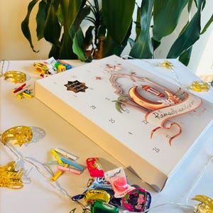 Pu&ograve; includere: Un calendario bianco del Ramadan con dettagli dorati, una luna crescente e un design a lanterna. Il calendario &egrave; decorato con piccole luci a forma di stella e luna e circondato da caramelle colorate e luci decorative.