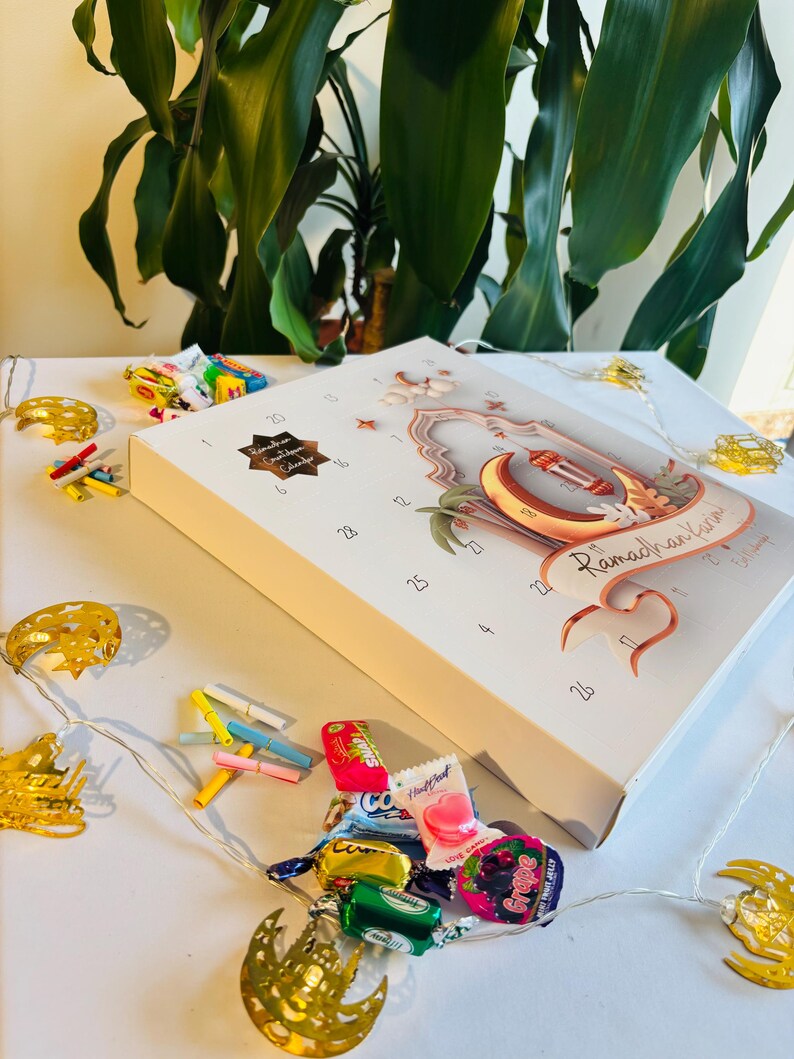 Pu&ograve; includere: Un calendario bianco del Ramadan con dettagli dorati, una luna crescente e un design a lanterna. Il calendario &egrave; decorato con piccole luci a forma di stella e luna e circondato da caramelle colorate e luci decorative.