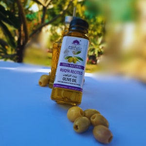 Puede incluir: Una botella de vidrio transparente de aceite de oliva con una etiqueta morada que dice "100% Natural Ruqya Recited Olive Oil" en &aacute;rabe e ingl&eacute;s. La botella est&aacute; sobre una superficie azul claro con aceitunas verdes esparcidas alrededor.