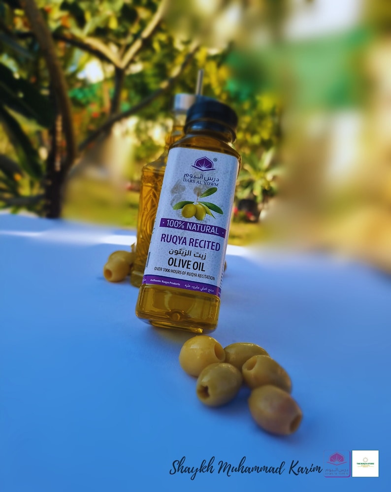 Puede incluir: Una botella de vidrio transparente de aceite de oliva con una etiqueta morada que dice "100% Natural Ruqya Recited Olive Oil" en &aacute;rabe e ingl&eacute;s. La botella est&aacute; sobre una superficie azul claro con aceitunas verdes esparcidas alrededor.