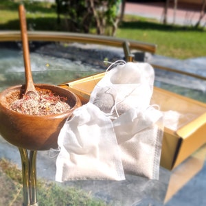 Pode incluir: Uma tigela de madeira cheia de uma mistura de especiarias marrons e vermelhas, com uma colher de madeira em cima. Dois sacos de musselina brancos estão em uma caixa dourada ao lado da tigela.