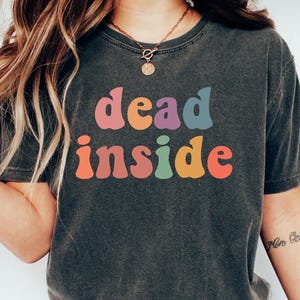 May include: Dark gray t-shirt with the words "dead inside" in colorful, retro-style lettering. The shirt is paired with blue jeans and white sneakers. The overall aesthetic is casual and relaxed.