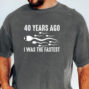 May include: A gray t-shirt with the text "40 YEARS AGO I WAS THE FASTEST" and a sperm cell graphic. The shirt is a casual style, with short sleeves and a relaxed fit. The text and graphic are in white.