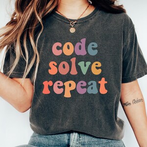 May include: Dark gray t-shirt with the words "code solve repeat" in colorful, retro-style lettering. The shirt is paired with blue jeans and white sneakers, creating a casual, everyday look.