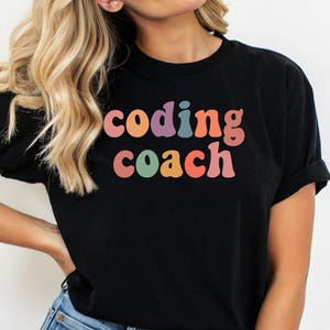 May include: Black t-shirt with the text "coding coach" in colorful, retro-style lettering. The shirt is a classic crew neck style and is made of a soft material. The shirt is paired with blue jeans.