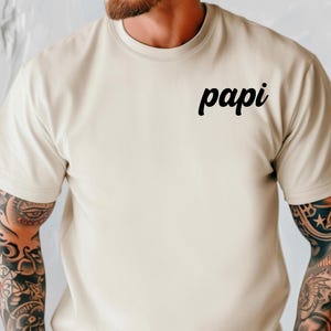 May include: A beige short-sleeved t-shirt with the word "papi" printed in black script on the upper chest. The person wearing the shirt has tattoos on both arms and is wearing a gold watch.