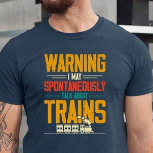 May include: A navy blue t-shirt with a vintage-style graphic that reads "WARNING I MAY SPONTANEOUSLY TALK ABOUT TRAINS". The graphic features a yellow, orange, and red color scheme and includes a small illustration of a train.