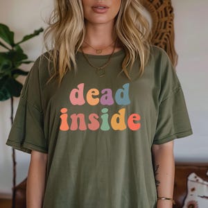 May include: Olive green t-shirt with the words "dead inside" in colorful, retro-style lettering. The text is in shades of pink, orange, blue, and yellow. The shirt has short sleeves and a relaxed fit.