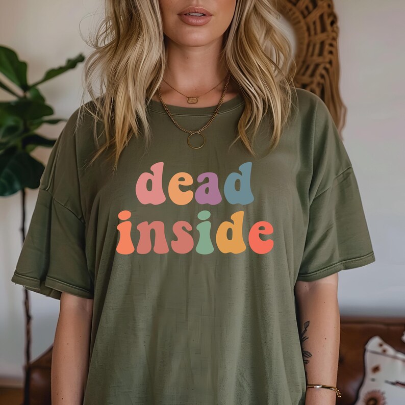 May include: Olive green t-shirt with the words "dead inside" in colorful, retro-style lettering. The text is in shades of pink, orange, blue, and yellow. The shirt has short sleeves and a relaxed fit.