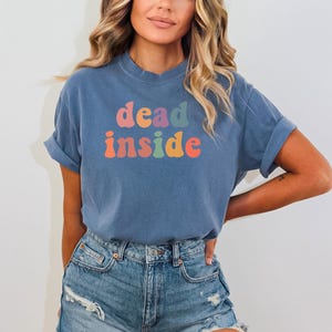 May include: A blue t-shirt with the words "dead inside" in colorful, retro-style lettering. The shirt is made of a soft material and is paired with denim shorts. The overall aesthetic is casual and comfortable.