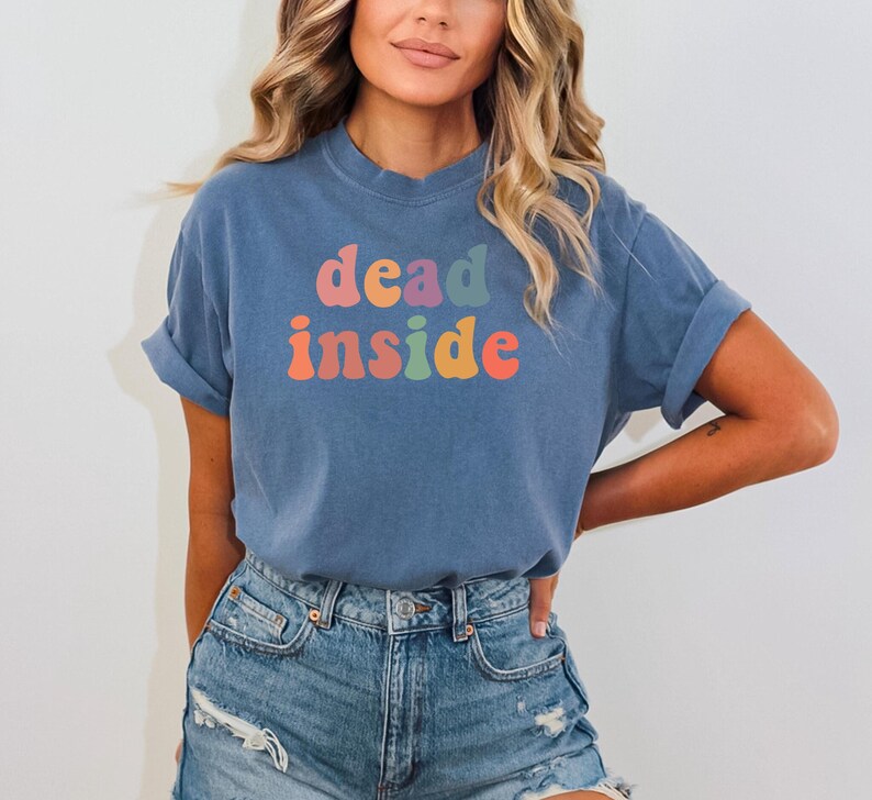 May include: A blue t-shirt with the words "dead inside" in colorful, retro-style lettering. The shirt is made of a soft material and is paired with denim shorts. The overall aesthetic is casual and comfortable.