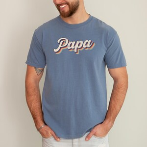 May include: A blue t-shirt with the word "Papa" in a retro, rainbow-colored font.