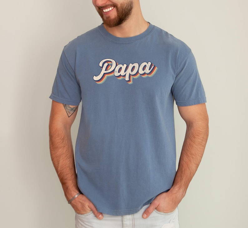 May include: A blue t-shirt with the word "Papa" in a retro, rainbow-colored font.