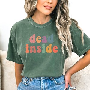 May include: Green t-shirt with the words "dead inside" in colorful, retro-style lettering. The shirt has a crew neck and short sleeves. The text is in shades of orange, pink, purple, and blue.