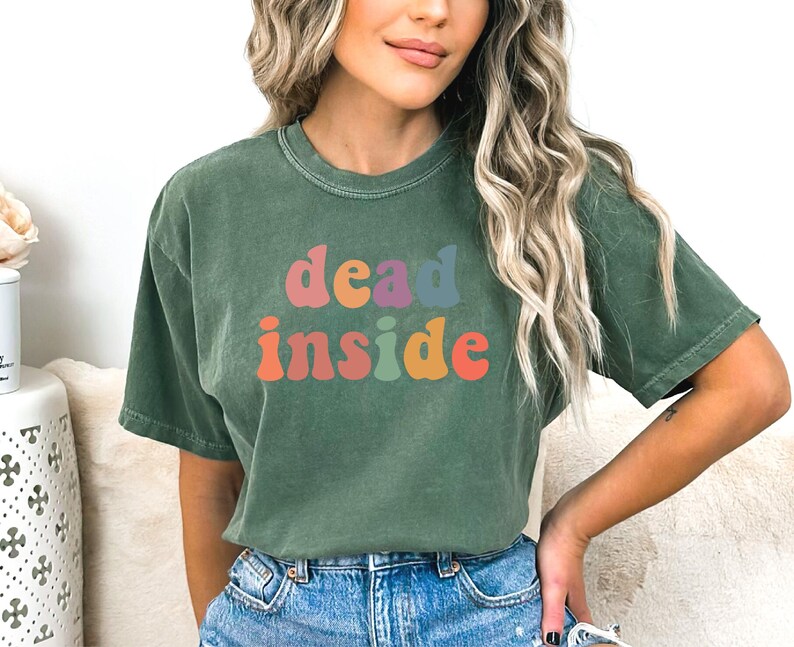 May include: Green t-shirt with the words "dead inside" in colorful, retro-style lettering. The shirt has a crew neck and short sleeves. The text is in shades of orange, pink, purple, and blue.