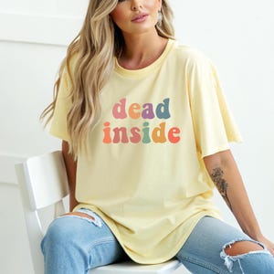 May include: A light yellow t-shirt with the words "dead inside" in colorful, retro-style lettering. The shirt has short sleeves and a relaxed fit. The model is wearing light blue jeans.