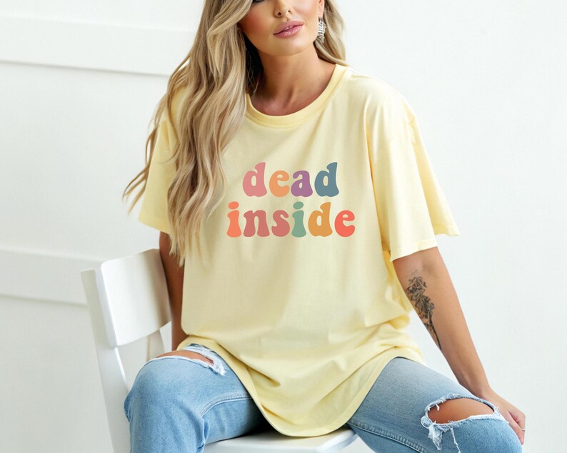 May include: A light yellow t-shirt with the words "dead inside" in colorful, retro-style lettering. The shirt has short sleeves and a relaxed fit. The model is wearing light blue jeans.