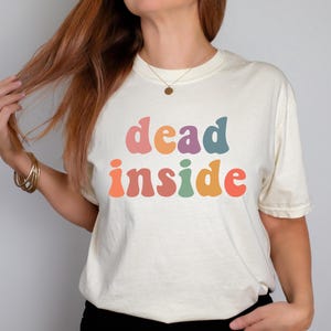 May include: Cream-colored t-shirt with the words "dead inside" in colorful, retro-style lettering. The text is in shades of pink, orange, purple, green, and red. The shirt has short sleeves and a crew neck.