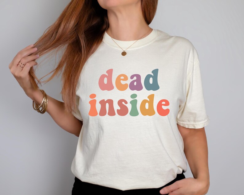 May include: Cream-colored t-shirt with the words "dead inside" in colorful, retro-style lettering. The text is in shades of pink, orange, purple, green, and red. The shirt has short sleeves and a crew neck.