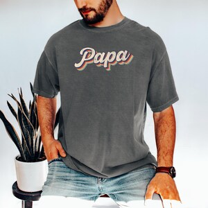 May include: A dark gray t-shirt with a retro-style graphic that says "Papa" in colorful letters.