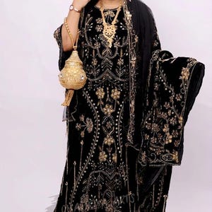 May include: Black velvet dress with intricate gold embroidery and beadwork. The dress features a long, flowing silhouette, a matching shawl, and a gold beaded purse. The ensemble includes gold jewelry and a black head covering.