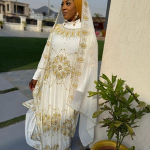 May include: A white gown with gold and pearl embroidery, worn with a white veil and a mustard yellow headscarf.