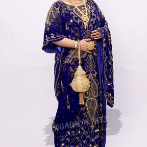 May include: A woman wearing a blue velvet kaftan with gold embroidery and a matching blue and gold dupatta. She is wearing gold jewelry and holding a gold clutch bag.