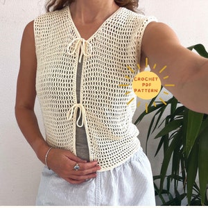 Op de afbeelding: Een wit gebreid vest met een strik sluiting. Het vest is mouwloos en heeft een losse pasvorm. De tekst "CROCHET PDF PATTERN" is zichtbaar in de afbeelding.