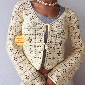 Könnte beinhalten: Eine gehäkelte cremefarbene Strickjacke mit Bindeband vorne und langen Ärmeln. Die Strickjacke hat ein zartes Lochmuster.