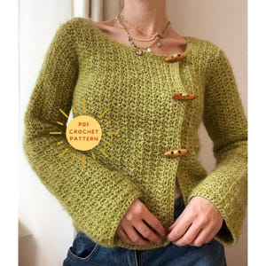 May include: A light green crochet cardigan with long sleeves and wooden toggle closures. The cardigan features a textured stitch pattern and a round neckline. The image includes the text "PDF Crochet Pattern" in a sunburst graphic.