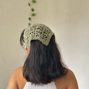 May include: A green crocheted headband with a delicate lace pattern. The headband is worn on a person with long, dark hair.