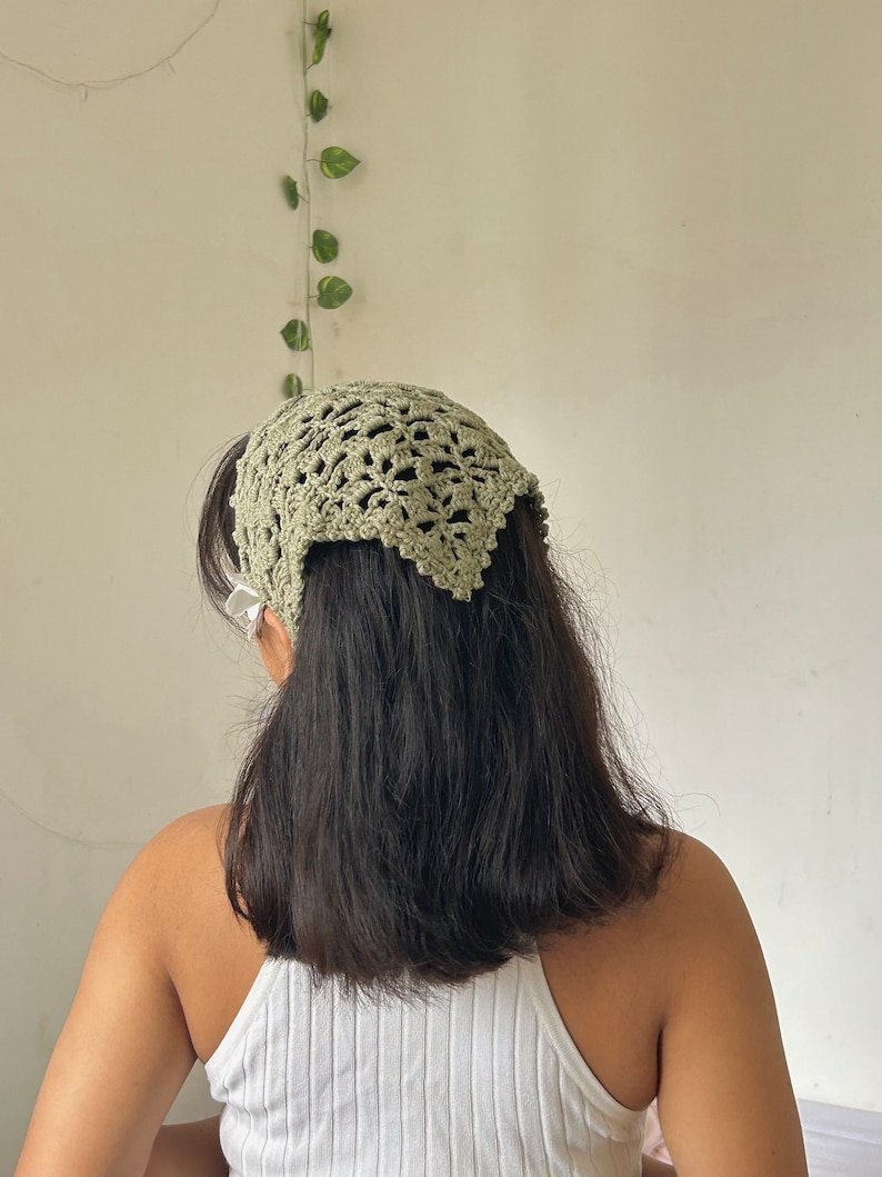 May include: A green crocheted headband with a delicate lace pattern. The headband is worn on a person with long, dark hair.