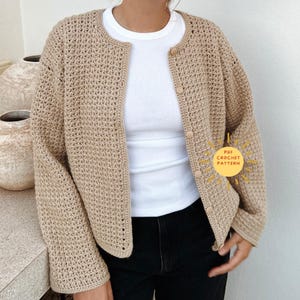Op de afbeelding: Een beige gehaakt vest met een ronde halslijn en houten knopen. Het open weefsel is zichtbaar en de mouwen zijn lang. Het vest wordt gedragen over een witte top en een zwarte broek. De afbeelding bevat een afbeelding met de tekst "PDF Crochet Pattern".