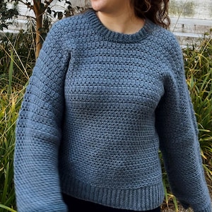 May include: A blue crocheted sweater with a textured stitch pattern. The sweater has a crew neckline and long sleeves.