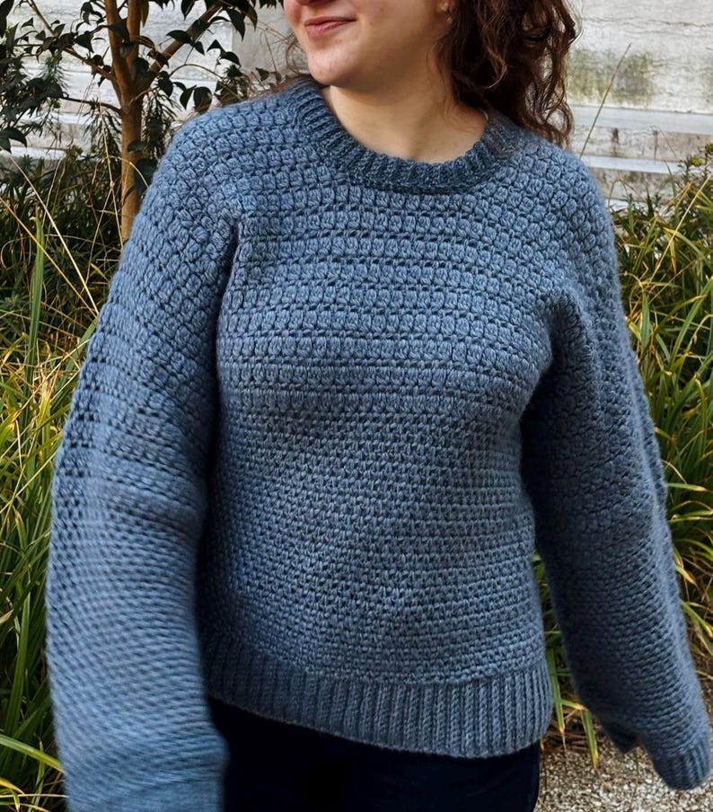 May include: A blue crocheted sweater with a textured stitch pattern. The sweater has a crew neckline and long sleeves.